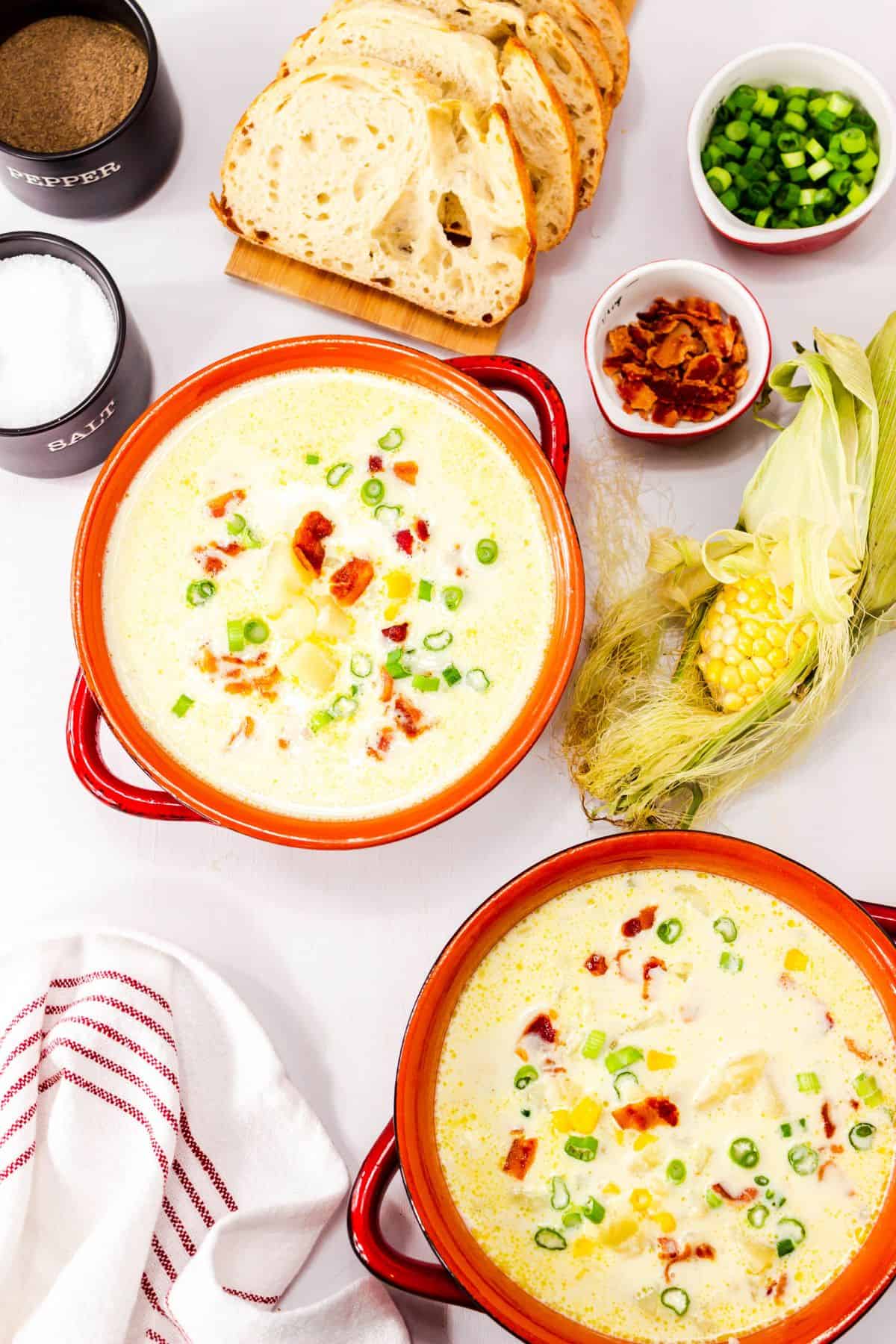 Two bowls  garnished with bacon and green onions, surrounded by sliced bread, salt, pepper,  and a bowl of chopped green onions.