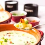 A spoonful of creamy dish with corn, potatoes, and green onions held above a bowl, with seasoning containers and more soup in the background.