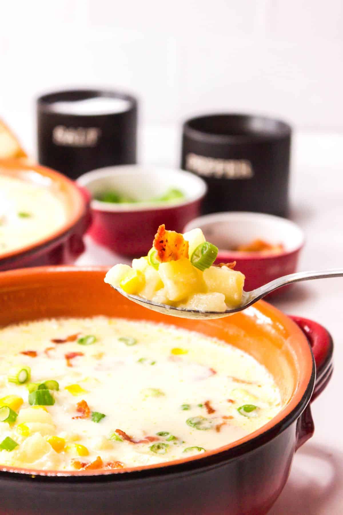 A spoonful of creamy dish with corn, potatoes, and green onions held above a bowl, with seasoning containers and more soup in the background.