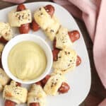 A plate of Little Smokies pigs in a blanket arranged in a circle surrounds a bowl of mustard dip, garnished with everything bagel seasoning, all elegantly placed on a wooden table adorned with a pink cloth.