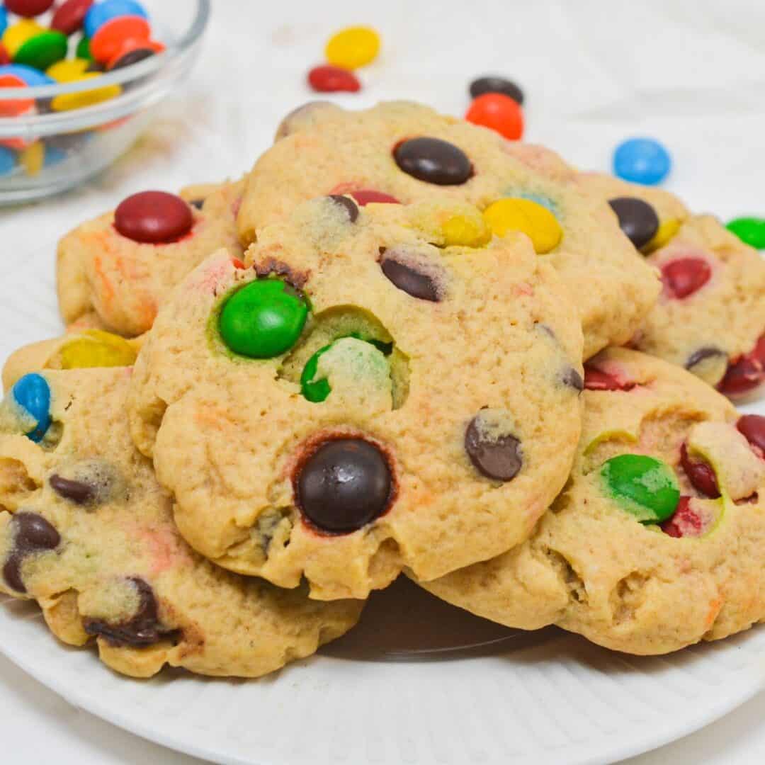 A plate of M&M Chocolate Chip Cookies topped with colorful candy-coated chocolates, with a bowl of similar candies in the background.