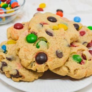 A plate of M&M Chocolate Chip Cookies topped with colorful candy-coated chocolates, with a bowl of similar candies in the background.