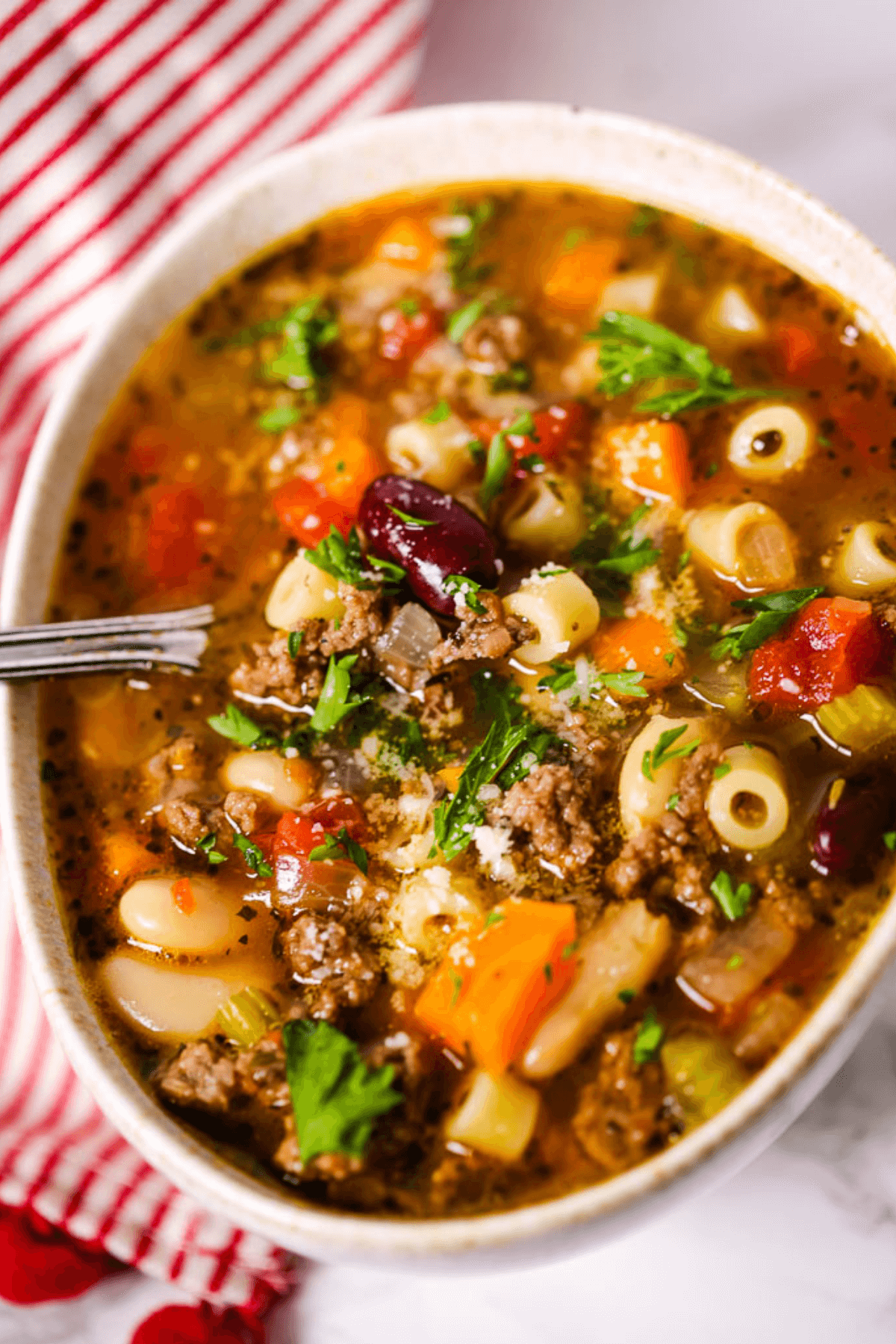 A bowl of hearty vegetable and beef soup with pasta, reminiscent of Olive Garden's Pasta Fagioli, garnished with parsley, rests beside a red and white striped cloth.