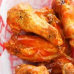 A basket of crispy, golden oven-baked buffalo wings coated in bright orange sauce sits invitingly on white paper.