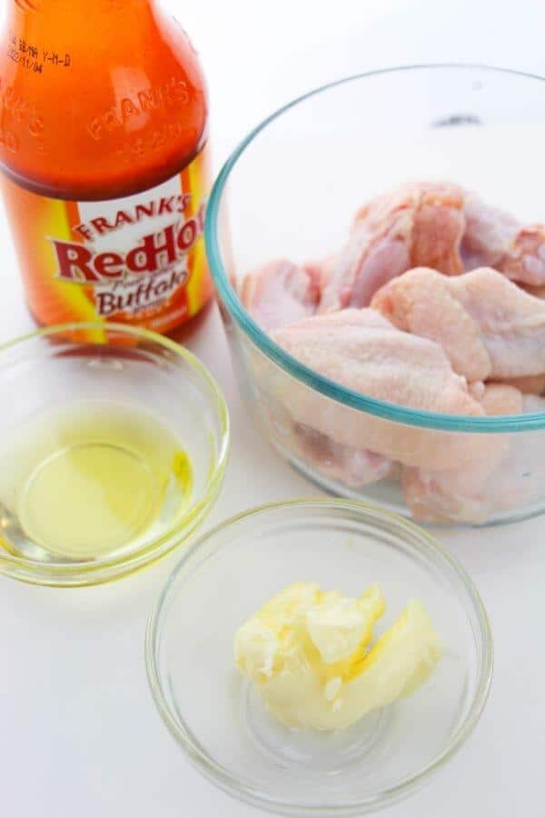A bottle of hot sauce, raw chicken wings in a bowl ready for oven-baked Buffalo wings, oil in a small dish, and butter in another small dish.