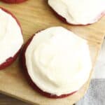 Four Red Velvet Cookies with Cream Cheese Frosting are arranged on a wooden board next to a white and gray cloth.