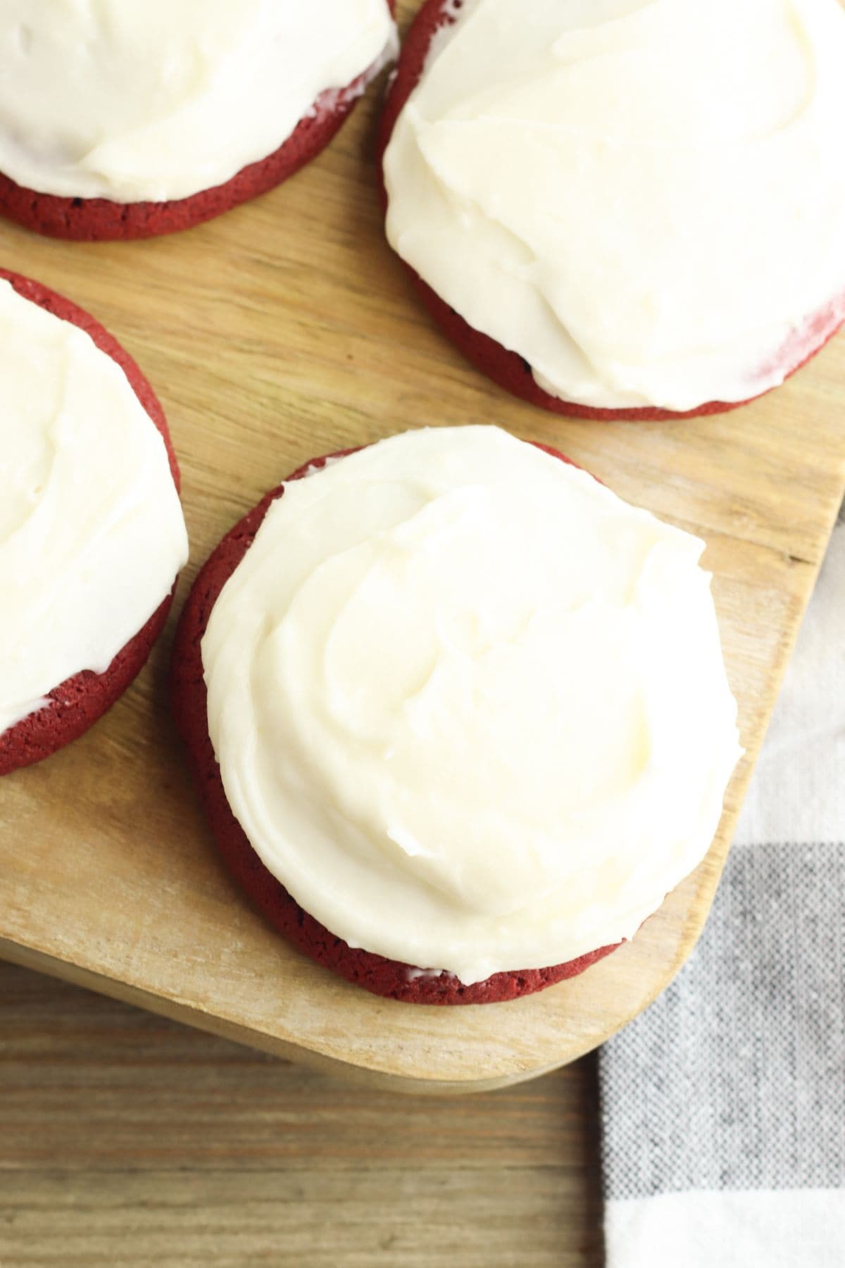Three Red Velvet Cookies with Cream Cheese Frosting are arranged on a wooden board, with part of a striped cloth visible in the corner.