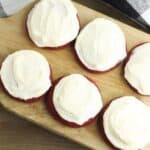 Six Red Velvet Cookies with Cream Cheese Frosting are arranged on a wooden board, with a black and white checkered cloth partially visible in the background.