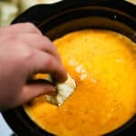 A hand dips a tortilla chip into a pot of melted, slow cooker cheese dip, reminiscent of rich pizza flavors.