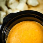 A black pot cradles a warm, orange soup reminiscent of slow cooker pizza dip, while slices of bread rest invitingly on a wooden board in the background.