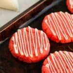 Three round Strawberry Cake Mix Cookies with white icing drizzled in diagonal lines sit on a dark baking sheet. A piping bag filled with icing is visible in the background.