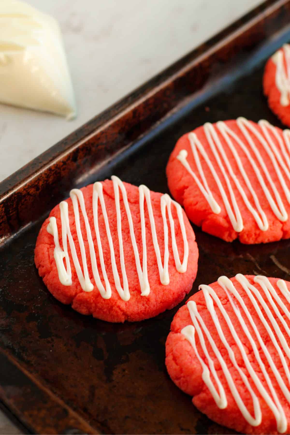 Three round Strawberry Cake Mix Cookies with white icing drizzled in diagonal lines sit on a dark baking sheet. A piping bag filled with icing is visible in the background.