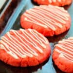 Four round, pink snacks with white icing drizzled in stripes, arranged on a dark baking sheet.
