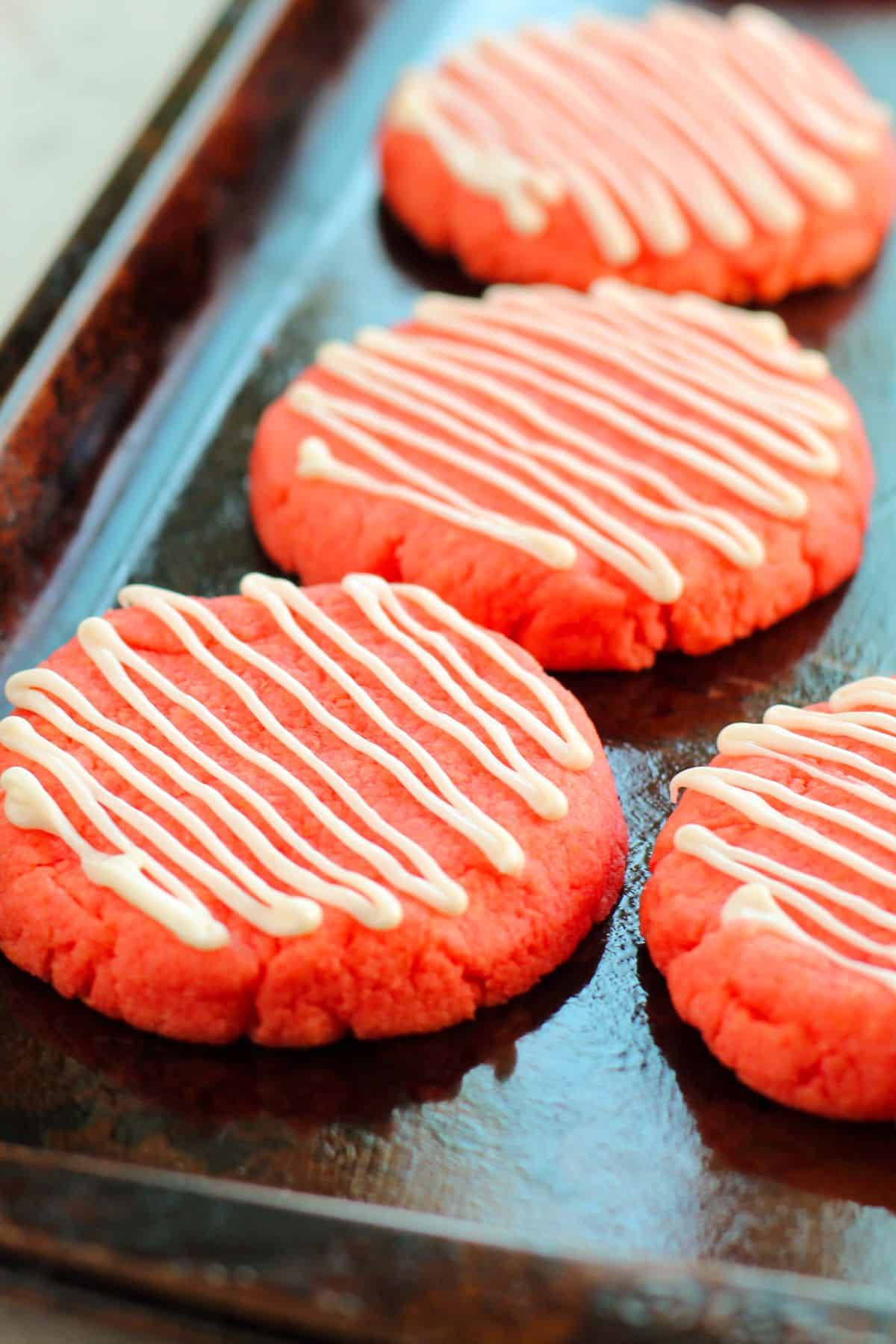 Four round, pink snacks with white icing drizzled in stripes, arranged on a dark baking sheet.