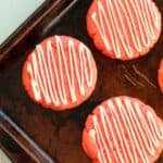 Strawberry Cake Mix Cookies with white zigzag icing rest on a dark metal baking sheet, their vibrant red color making them look as delightful as they taste.