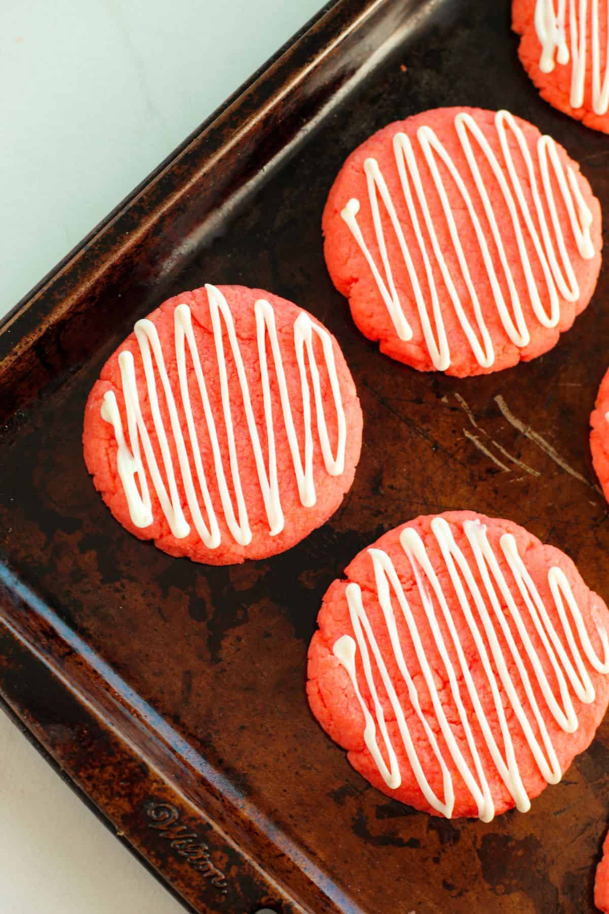 Strawberry Cake Mix Cookies with white zigzag icing rest on a dark metal baking sheet, their vibrant red color making them look as delightful as they taste.