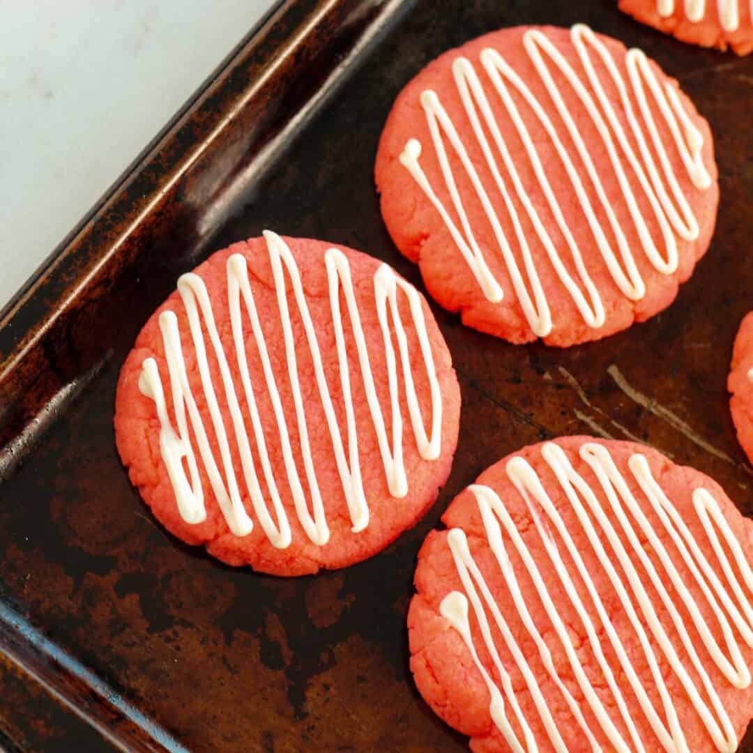 Strawberry Cake Mix Cookies with white icing in a zigzag pattern are arranged on a dark baking sheet, creating a colorful and inviting treat.