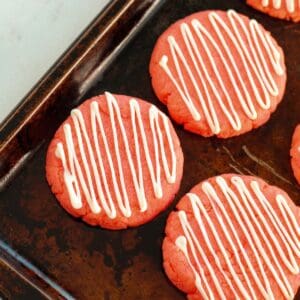 Strawberry Cake Mix Cookies with white icing in a zigzag pattern are arranged on a dark baking sheet, creating a colorful and inviting treat.