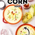 Two bowls of creamy Lighter Corn Chowder topped with green onions and bacon bits, surrounded by corn, bread, and small bowls of salt, pepper, and toppings on a white surface.