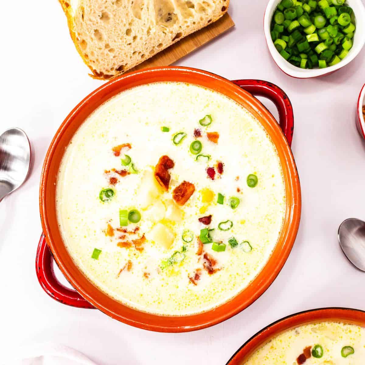 A bowl of Lighter Corn Chowder topped with chopped green onions and bacon, served with a slice of bread and a bowl of green onions on the side.