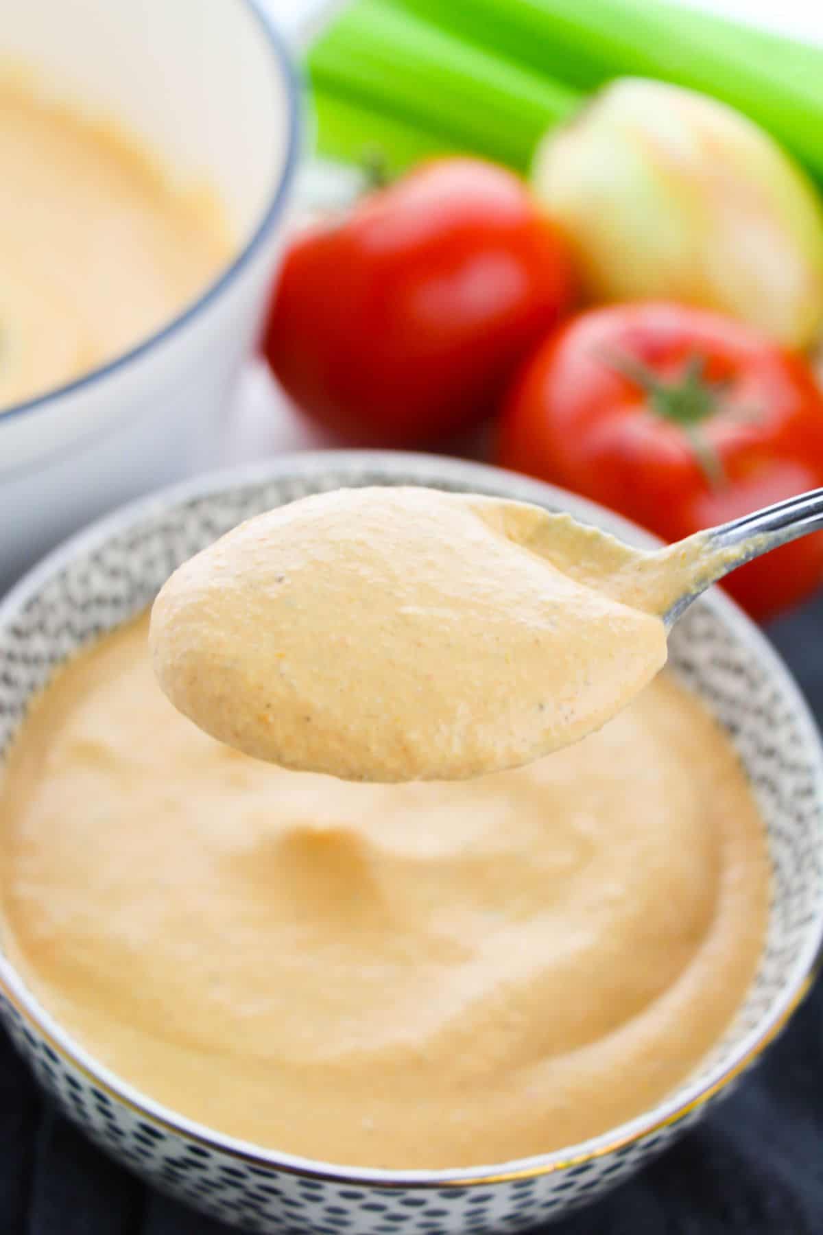 A spoon holding thick, creamy orange Tomato Basil Bisque above a bowl, with tomatoes, celery, and an onion in the background.