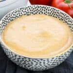 A bowl of creamy orange Tomato Basil Bisque sits in a patterned dish, with tomatoes and an onion in the background.