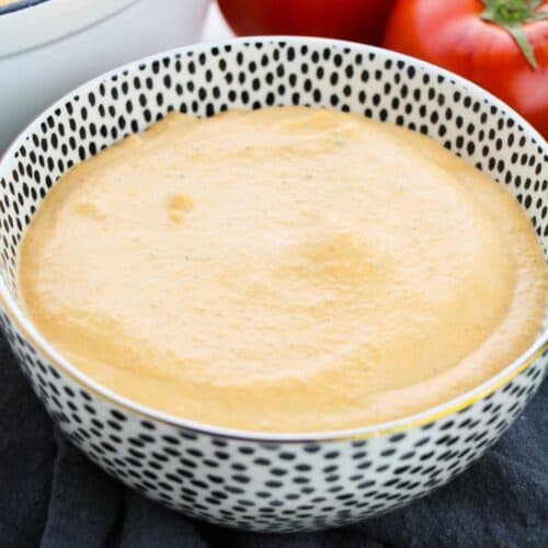 A bowl of creamy orange Tomato Basil Bisque sits in a patterned dish, with tomatoes and an onion in the background.