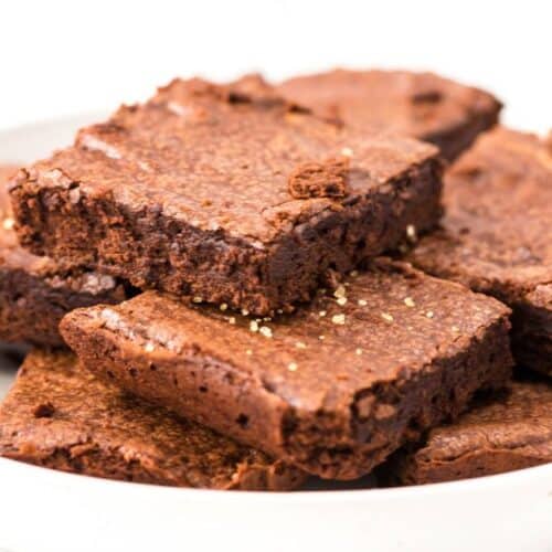 A white plate with several square Triple Chocolate Brownies stacked on it, with a few gold sprinkles visible on top of one brownie.