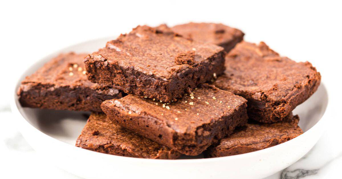 A white plate with several square Triple Chocolate Brownies stacked on it, with a few gold sprinkles visible on top of one brownie.