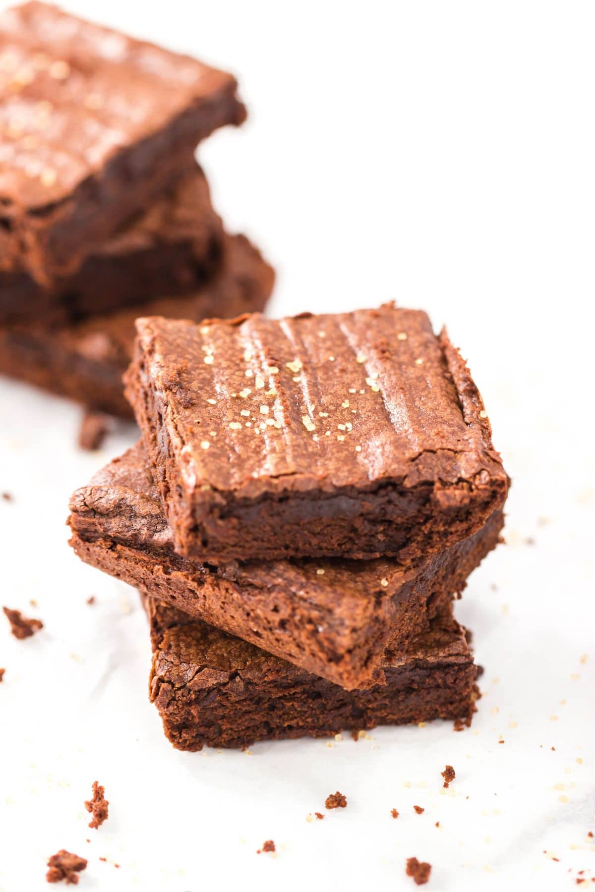 A stack of three Triple Chocolate Brownies sits on a white surface, with another stack of brownies blurred in the background.