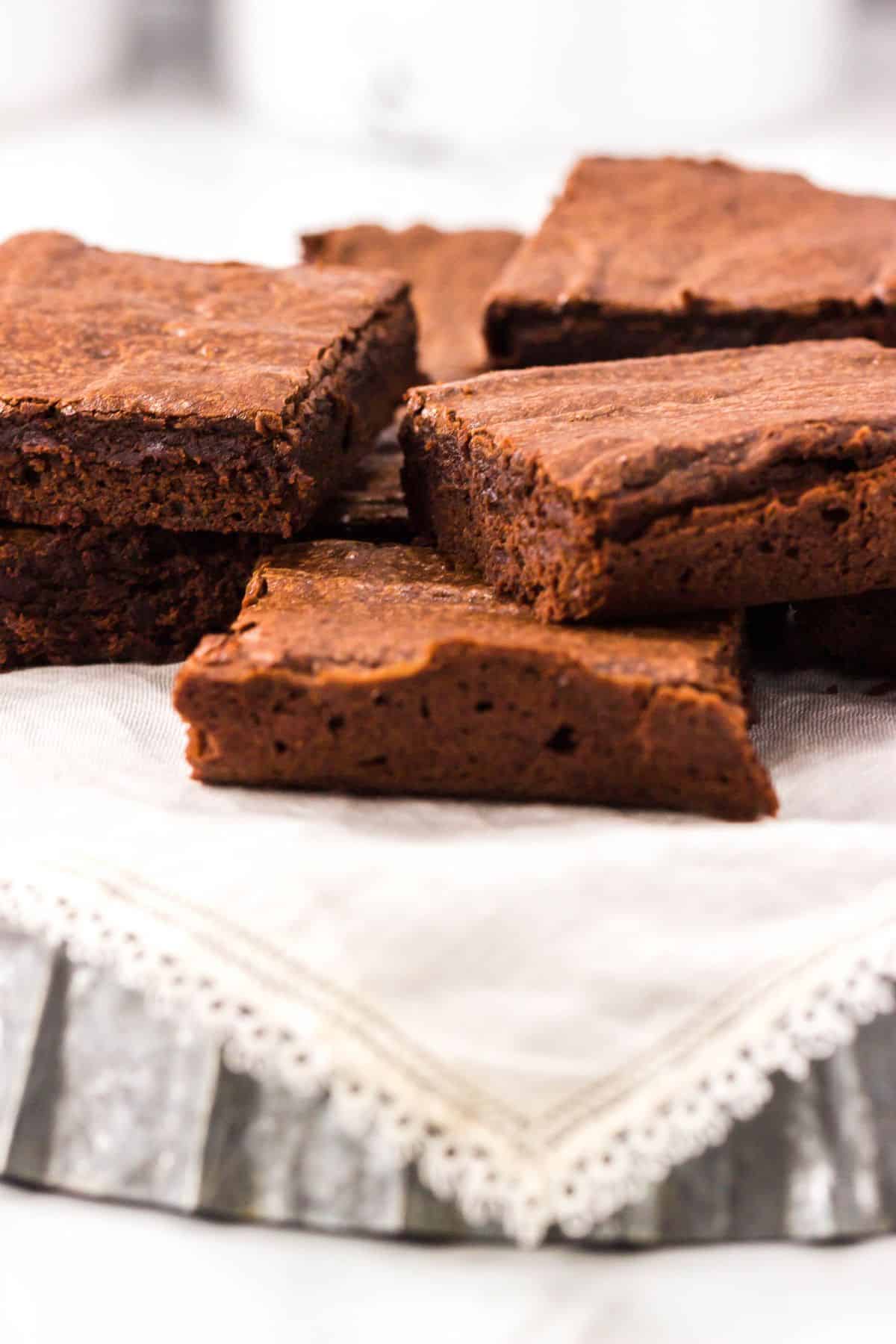 Several Triple Chocolate Brownies are stacked on a white cloth with lace trim, displayed on a metal tray.