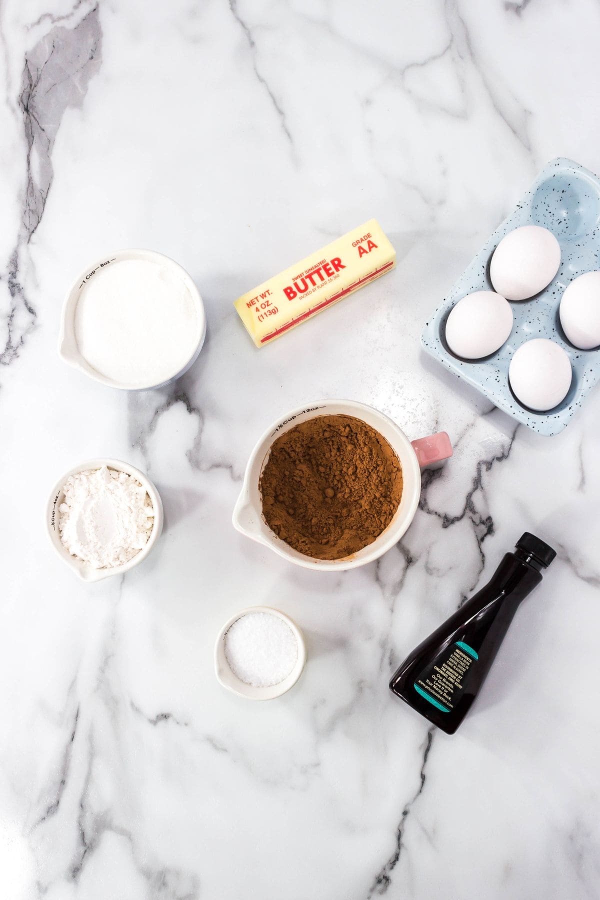 Baking ingredients for Triple Chocolate Brownies on a marble surface: a stick of butter, four eggs in a blue dish, bowls of sugar, cocoa powder, flour, salt, and a bottle of vanilla extract.
