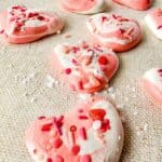 Heart-shaped pink and white Meringue Cookies with colorful sprinkles are scattered on a textured beige surface.