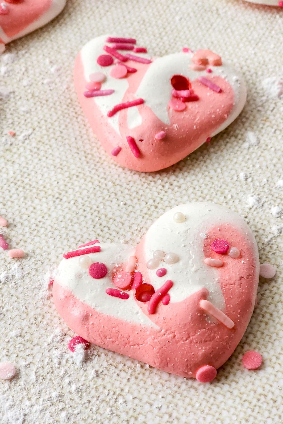 Heart-shaped pink and white Meringue Cookies adorned with assorted pink and red sprinkles, displayed on a textured surface.