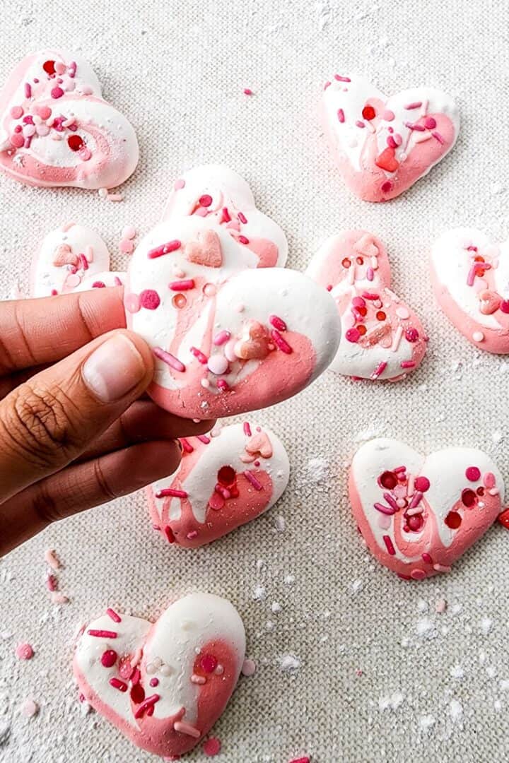 A hand holds a heart-shaped, pink and white meringue cookie decorated with pink and red sprinkles; several similar meringue cookies are scattered on a light surface nearby.