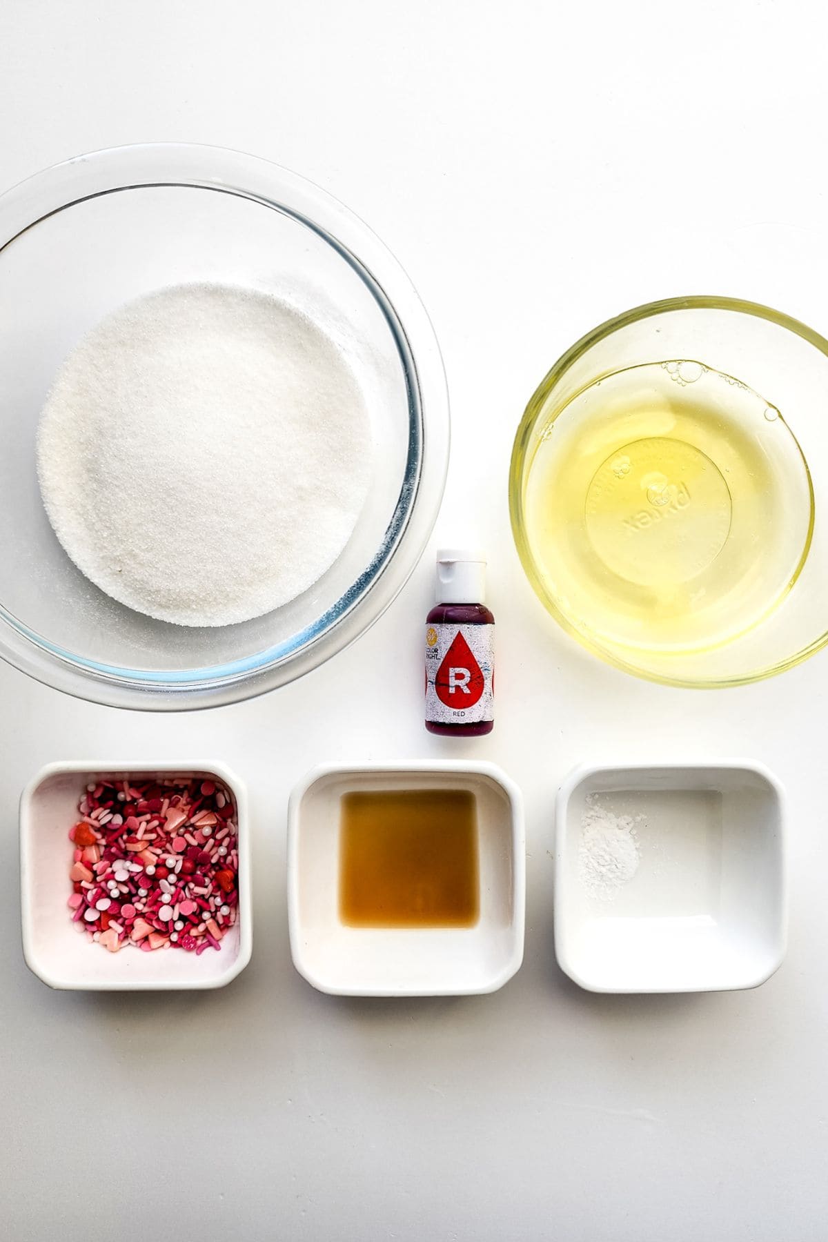 A glass bowl of sugar, a bowl of egg whites for Meringue Cookies, red food coloring, heart-shaped sprinkles, vanilla extract, and cream of tartar on a white surface.