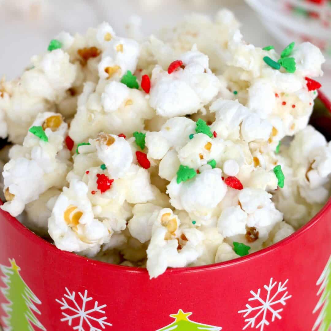 A red bowl decorated with green and yellow Christmas trees is filled with chocolate covered popcorn, coated in white topping and sprinkled with red and green festive sprinkles.