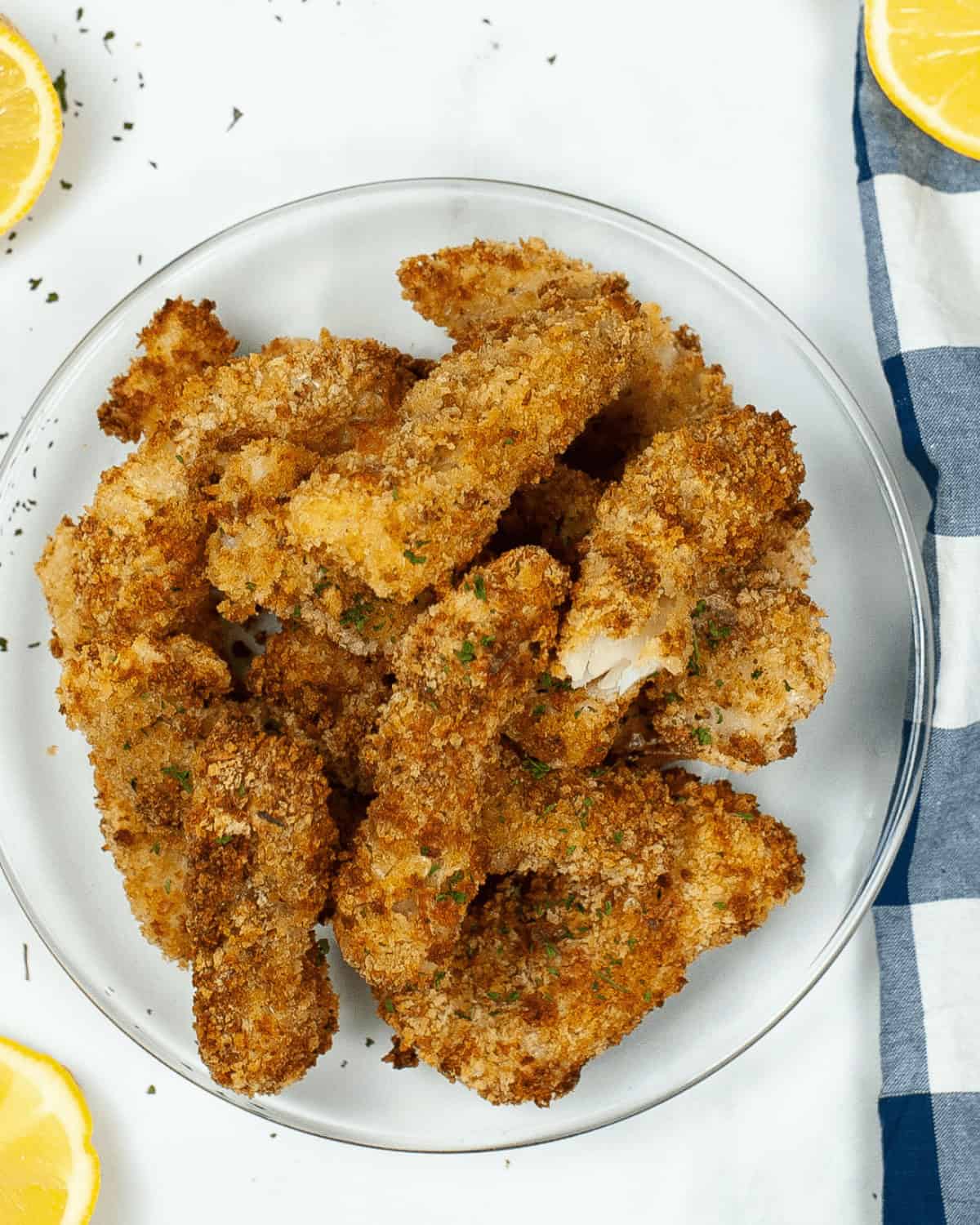 A plate of crispy, air fryer fish sticks garnished with parsley, accompanied by lemon wedges and a striped napkin.