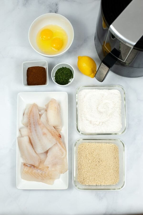 Ingredients for cooking Air Fryer Fish Sticks arranged on a kitchen countertop with fish, flour, breadcrumbs, and additional seasoning items.
