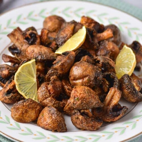 Plate of roasted Air Fryer Mushrooms garnished with three lime wedges, placed on a patterned plate atop a green textured cloth. Forks are visible in the blurred background.