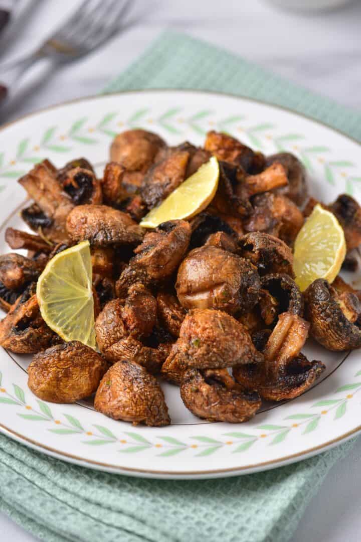Plate of roasted Air Fryer Mushrooms garnished with three lime wedges, placed on a patterned plate atop a green textured cloth. Forks are visible in the blurred background.