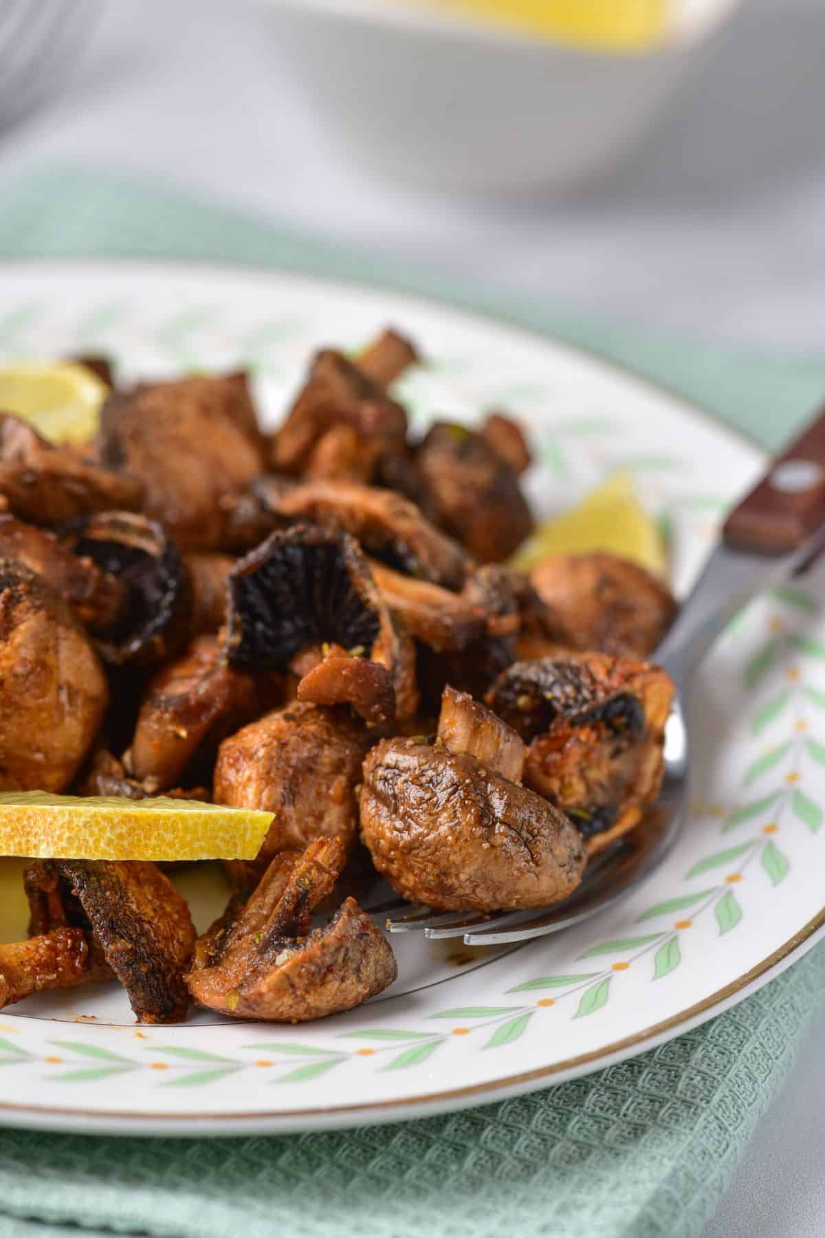 A plate of sautéed mushrooms, prepared Air Fryer Mushrooms style, with lemon wedges, served on a patterned dish with a fork resting on the side.
