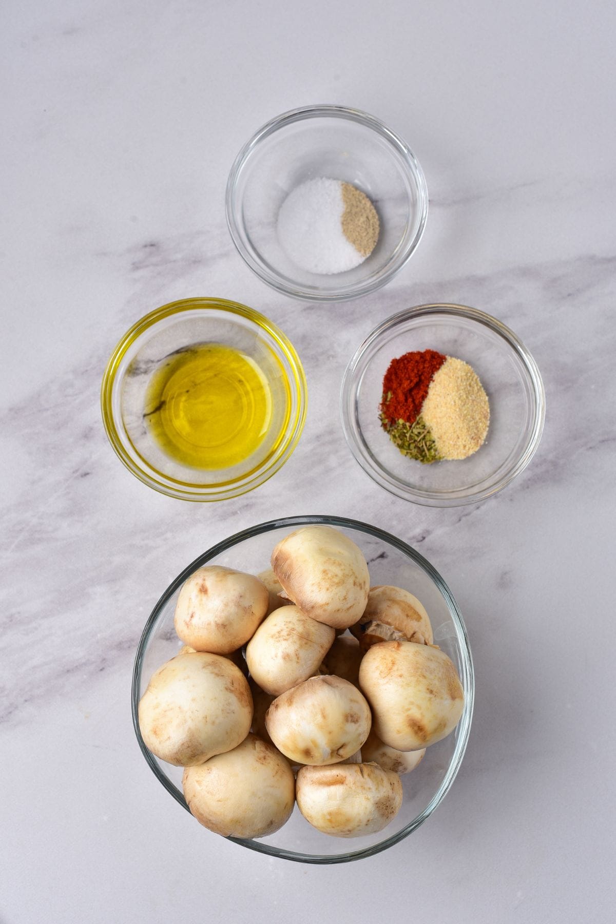 A bowl of whole white mushrooms, a bowl of olive oil, a bowl of salt, and a bowl of mixed spices on a white marble surface—perfect ingredients for delicious Air Fryer Mushrooms.
