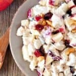 A plate with diced apples, dried cranberries, nuts, and creamy dressing sits on a wooden table next to a red apple and a wooden spoon.