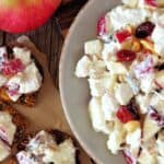A bowl of Apple Cranberry Salad with chopped apples, celery, nuts, and dried fruit in a creamy dressing, next to seeded crackers topped with the salad and a whole red apple.