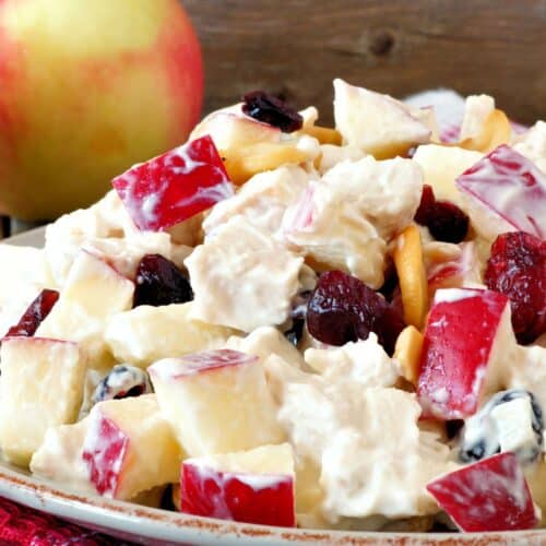 A plate of Apple Cranberry Salad with chopped red apples, celery, nuts, and dried cranberries in a creamy dressing, set on a red napkin with an apple in the background.