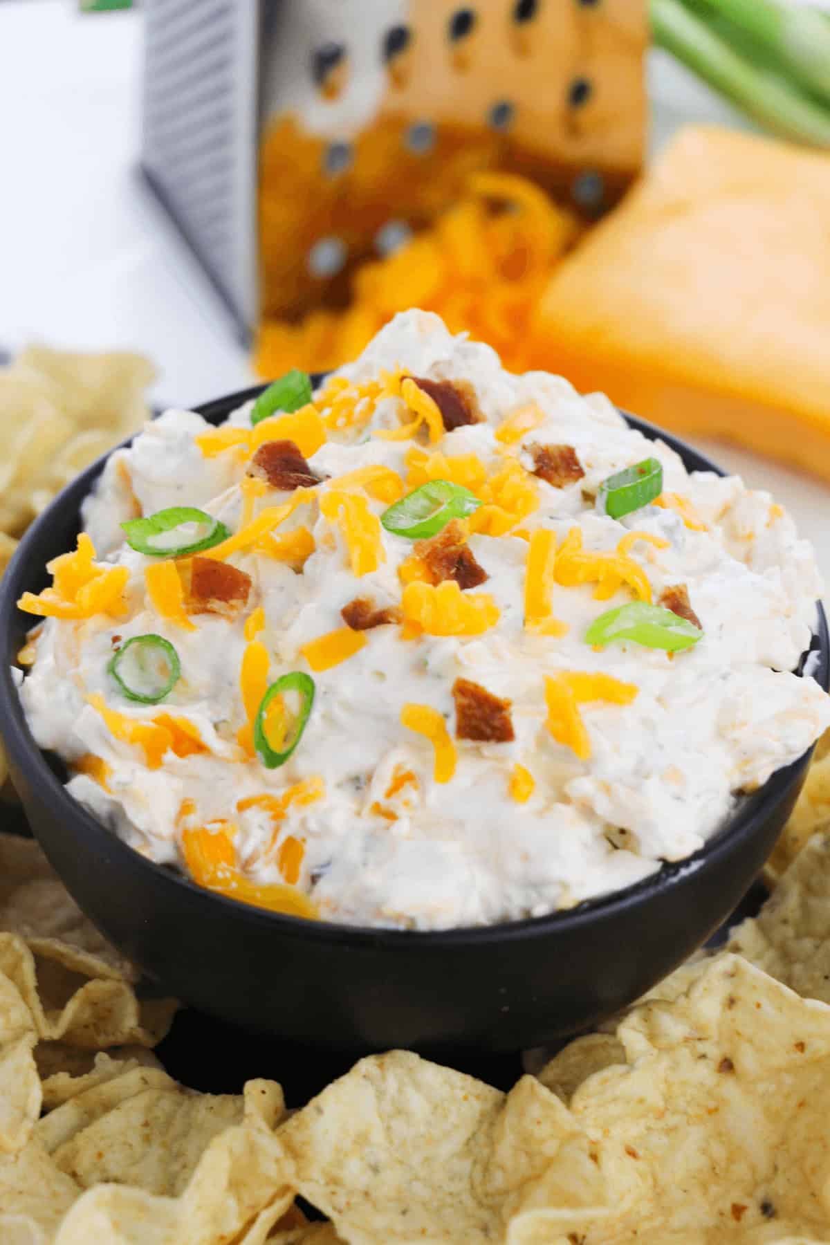 A bowl of creamy Bacon Cheddar Ranch Dip topped with shredded cheese, crispy bacon bits, and sliced green onions, surrounded by crunchy tortilla chips.