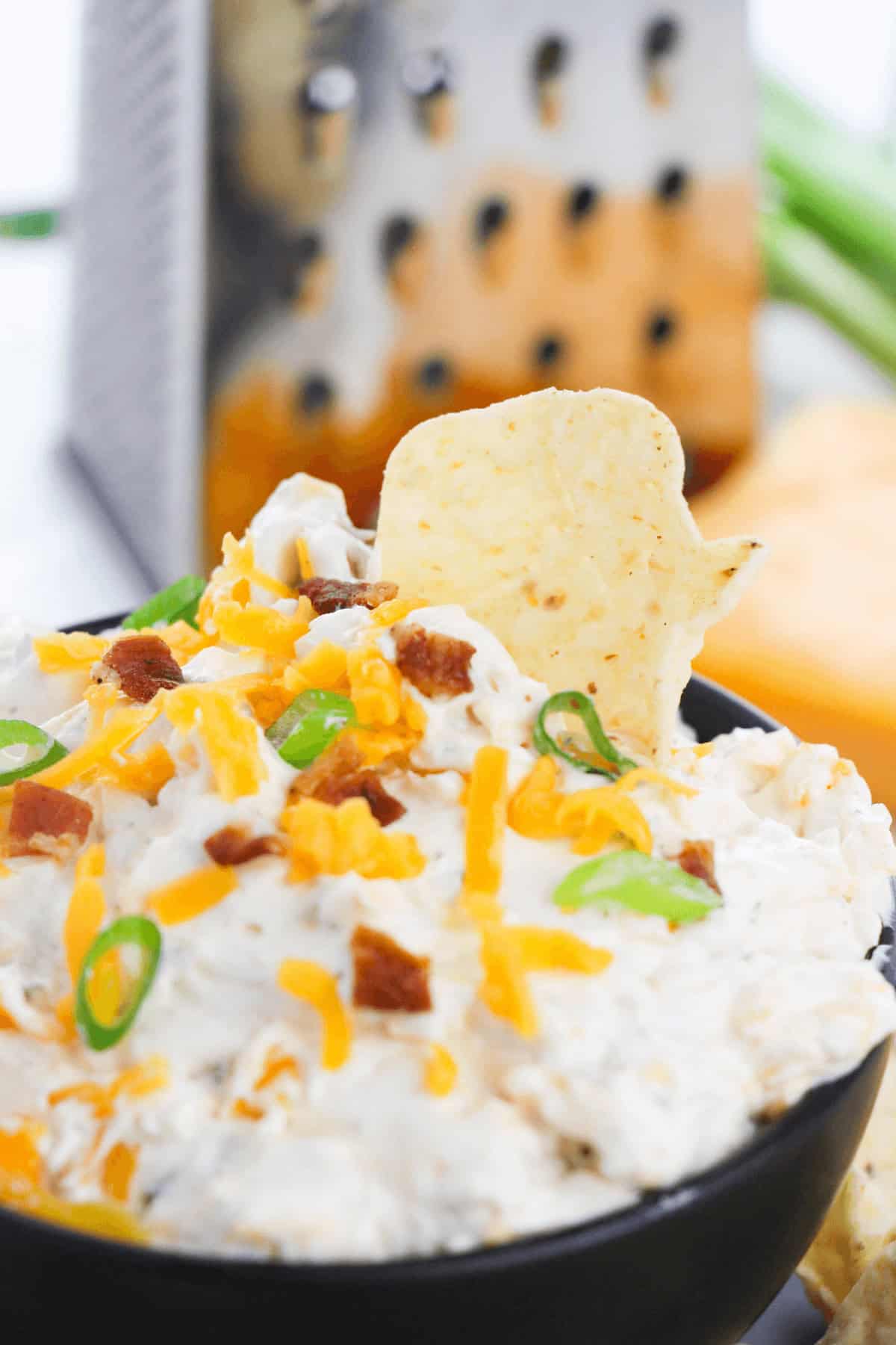 A close-up of a bowl of appetizer, topped with shredded cheese, bacon bits, and green onion slices. A chip is sticking out of the creamy dip. A cheese grater and ingredients are blurred in the background.