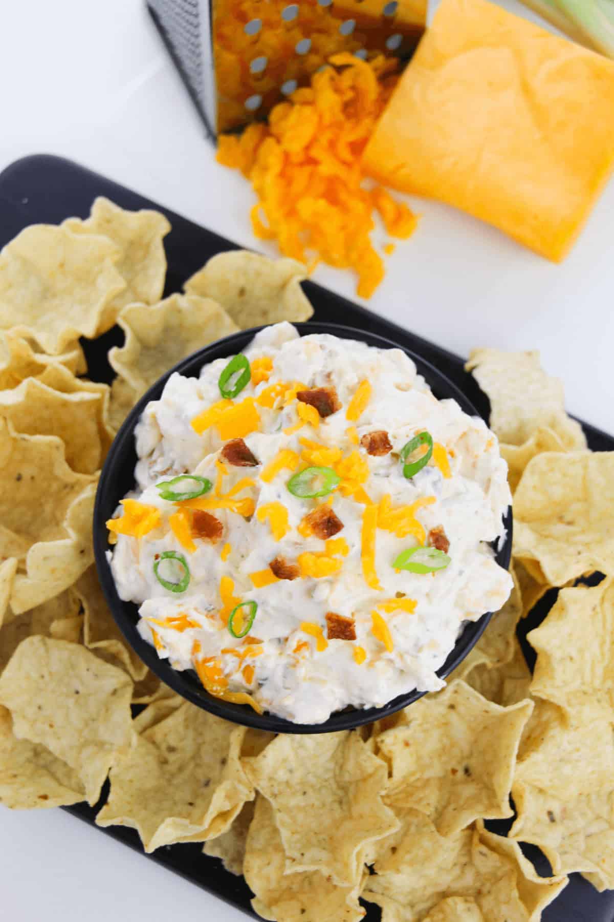 A bowl of Bacon Cheddar Ranch Dip garnished with cheese, green onions, and bacon bits is surrounded by tortilla chips. A block of cheese and grater are in the background.