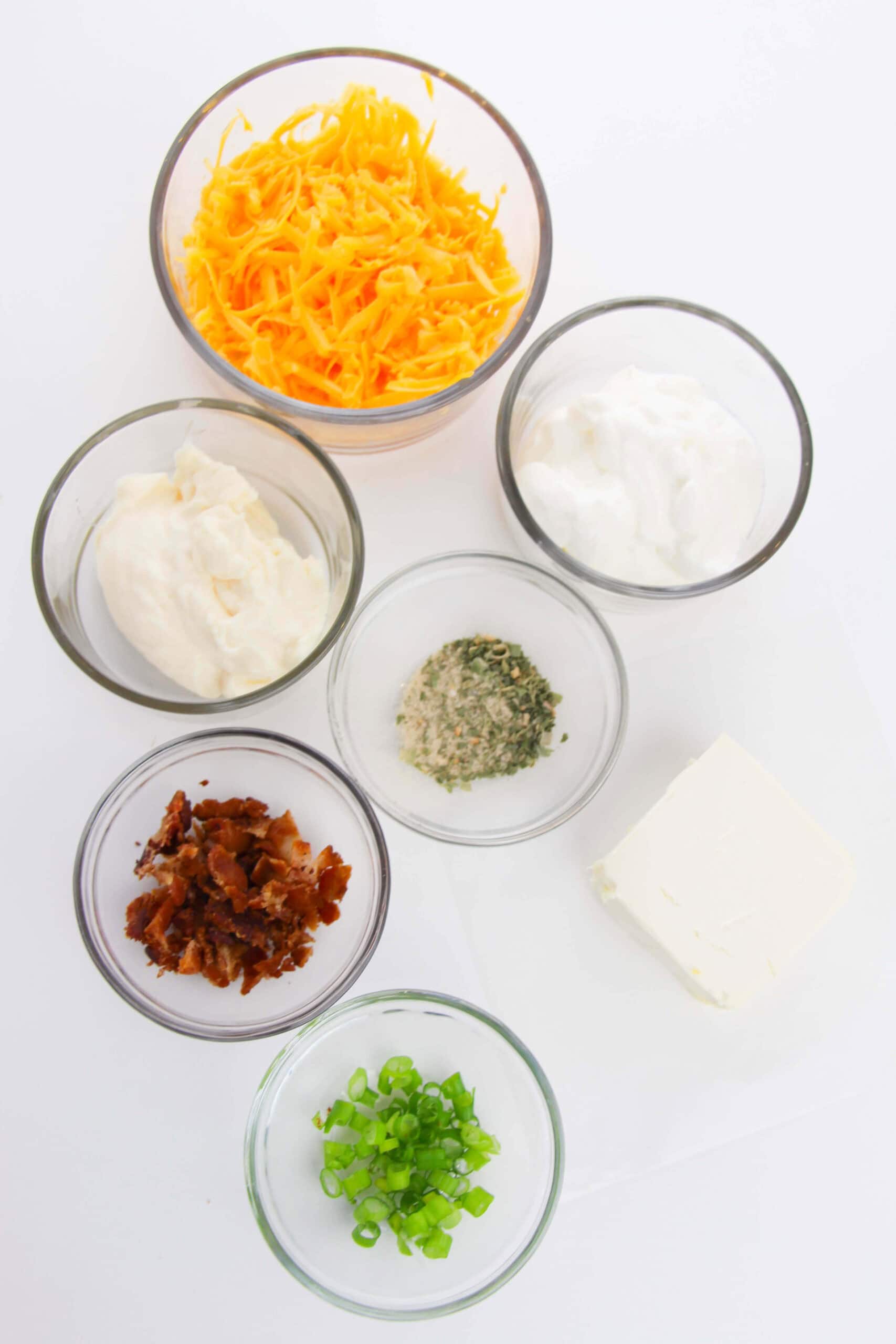 Glass bowls display the ingredients for a delectable Bacon Cheddar Ranch Dip: shredded cheese, sour cream, mayonnaise, crumbled bacon, chopped green onions, ranch seasoning, and a block of cream cheese on a pristine white surface.